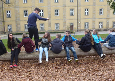 Jugendliche sitzen auf einem Baumstamm. Ein Teilnehmer versucht über diese hinwegzusteigen.