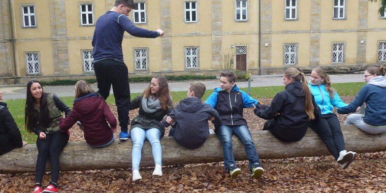 Jugendliche sitzen auf einem Baumstamm. Ein Teilnehmer versucht über diese hinwegzusteigen.