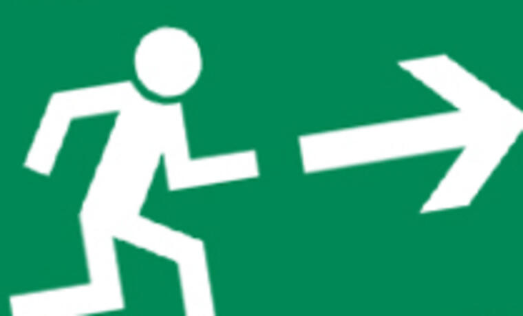 Grünes Schild mit weißen Strichmänchen. Dieses folgt einem weißen Pfeil zu einer Tür. Fluchttürschild.