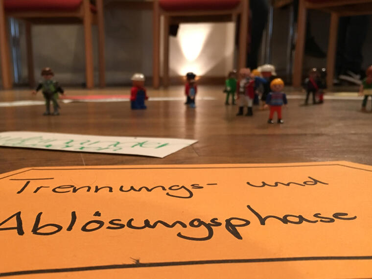 Vordergrund ein Schild mit der Aufschrift "Trennungs- und Ablösungsphase" im Hintergrund finden sich Playmobilfiguren